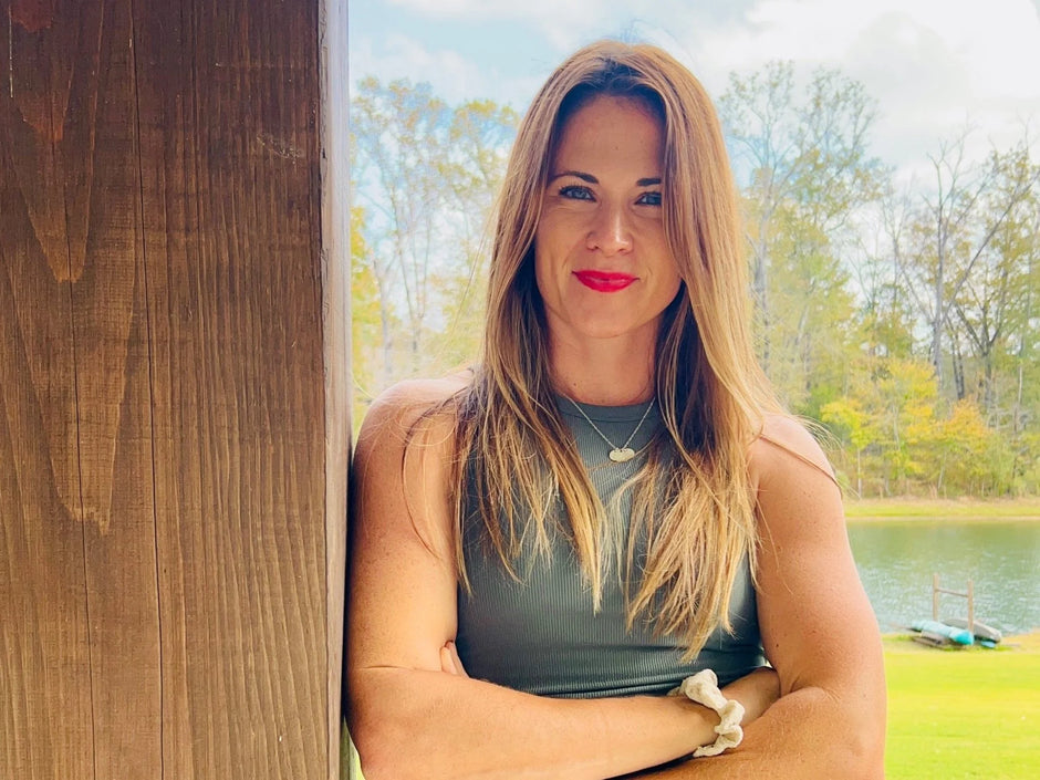 Woman with long blonde hair and red lipstick standing by a wooden structure with a scenic background of trees and water.